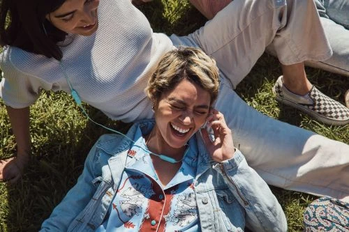 Two women relaxing on green grass, one laughing with earbuds, wearing a light blue denim jacket and printed shirt; the other with a white sweater.