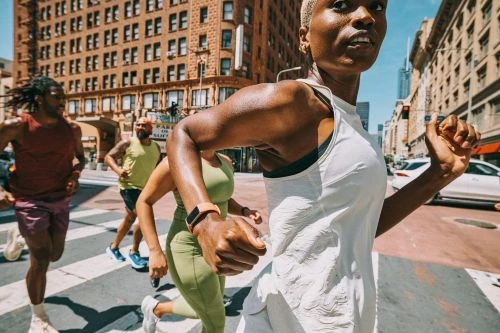 Four adults run across a crosswalk in an urban setting. The lead runner is a woman with short blonde hair wearing a white tank top.