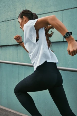 Woman in white t-shirt and black leggings running outdoors against a teal wall, wearing a smartwatch.