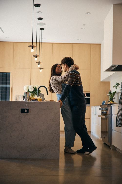 A couple embraces in a modern kitchen with warm lighting, featuring a marble island, wooden cabinets, and pendant lights.