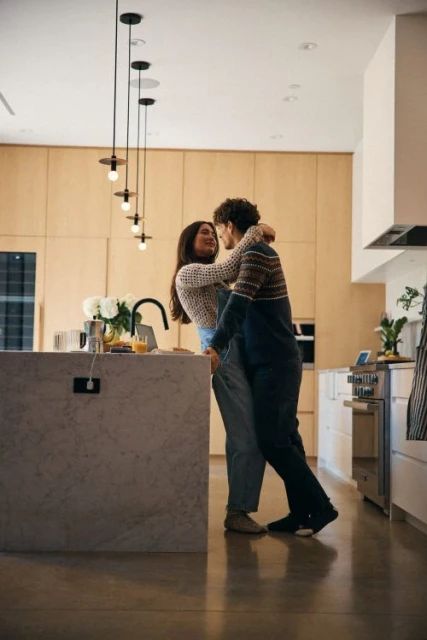 A couple embraces in a modern kitchen with warm lighting, featuring a marble island, wooden cabinets, and pendant lights.