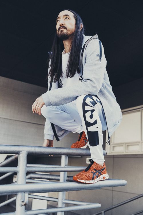 Steve Aoki poses in a grey hoodie, white pants, and orange sneakers, crouching outdoors on a metal railing with a serious expression.