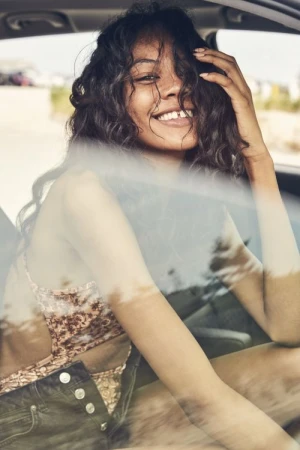 Smiling woman with dark curly hair sitting inside a car looking through the window by photographer Jeff Vallee.