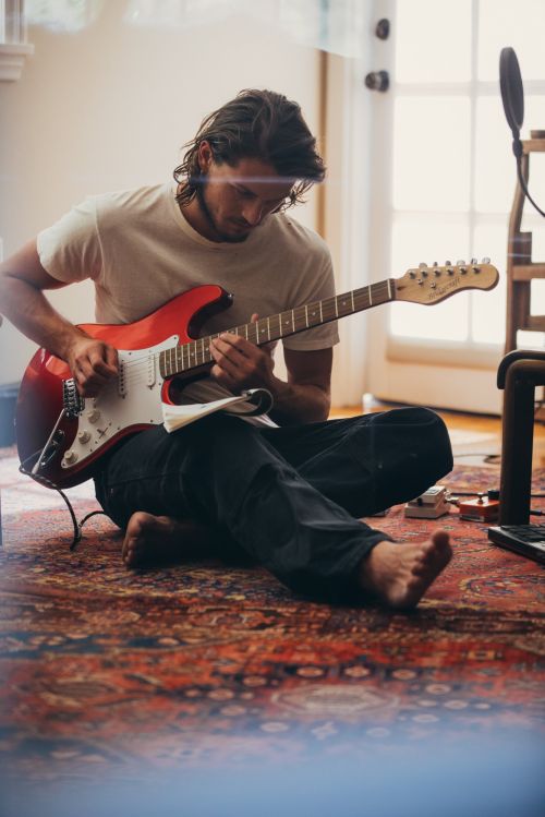 Man sits cross-legged on an ornate rug indoors, playing a red electric guitar with sheet music in a sunlit room, recording music.