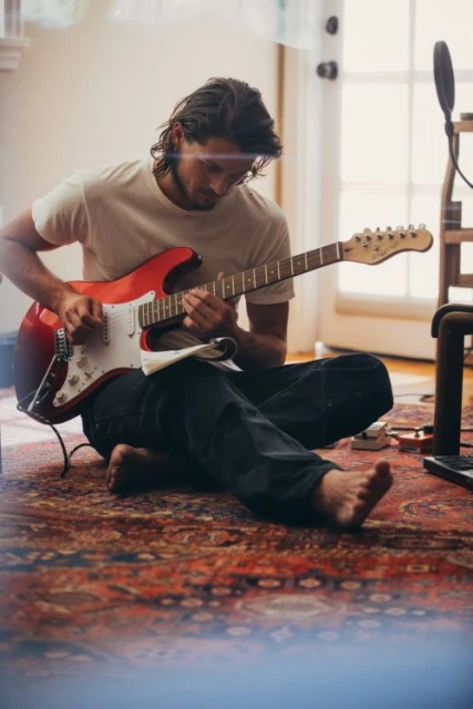 Man sits cross-legged on an ornate rug indoors, playing a red electric guitar with sheet music in a sunlit room, recording music.