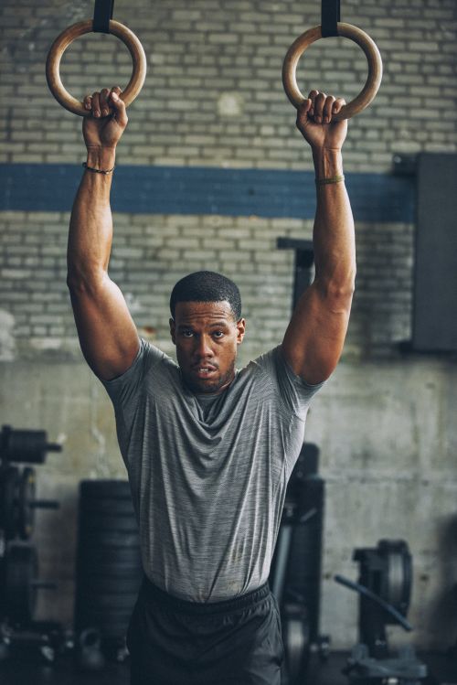 Muscular man in gray t-shirt hangs from gymnastic rings in a fitness center, flexing arms and looking at the camera with a serious expression.