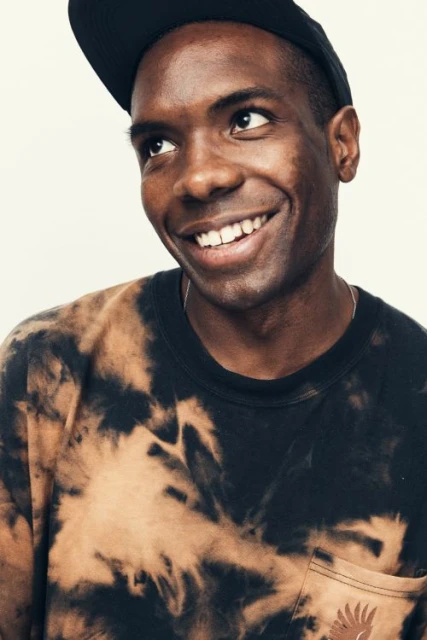 Portrait of a smiling Black man wearing a black tie-dye t-shirt and a baseball cap against a light background, head and shoulders.