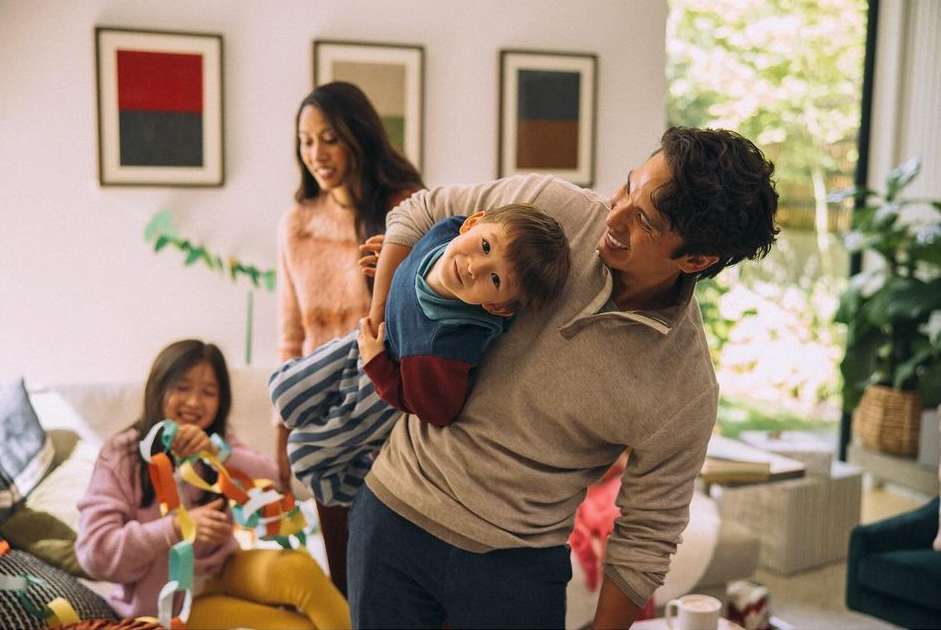 Family fun indoors with dad holding son, two girls creating paper chain, mother standing near window, and natural light.