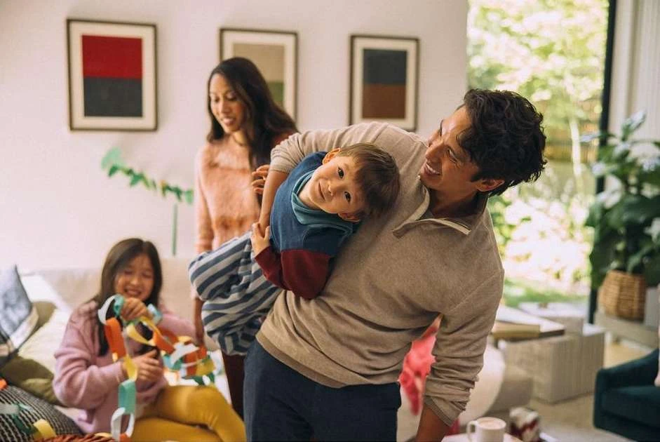 Family fun indoors with dad holding son, two girls creating paper chain, mother standing near window, and natural light.