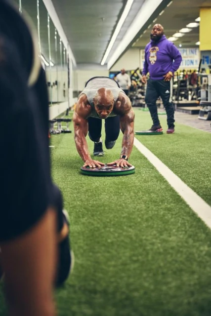 A muscular man is exercising in a gym in a plank position on a plate with another man watching him closely in the background.