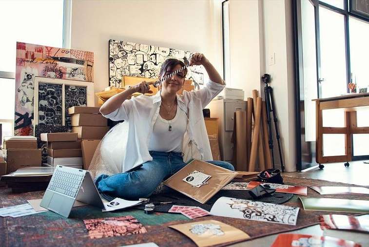 Adult female artist sits on the floor surrounded by art supplies and artworks in an indoor studio setting with natural lighting.