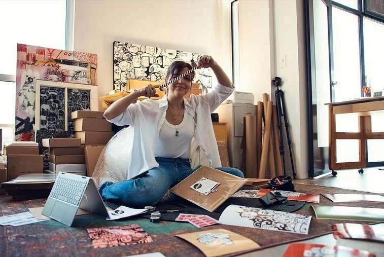 Adult female artist sits on the floor surrounded by art supplies and artworks in an indoor studio setting with natural lighting.