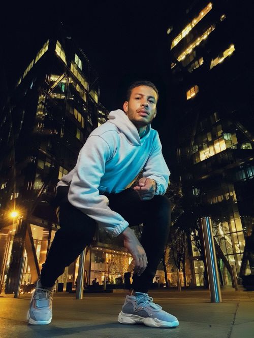Young man in hoodie and sneakers crouches on city street at night, illuminated by streetlights with office buildings in the background.