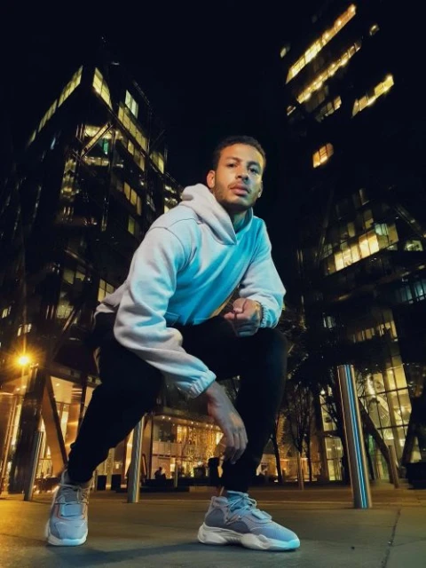 Young man in hoodie and sneakers crouches on city street at night, illuminated by streetlights with office buildings in the background.
