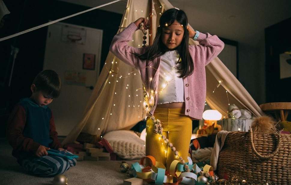 A young girl decorates a tent with string lights and wooden beads, while a young boy plays with blocks nearby indoors.