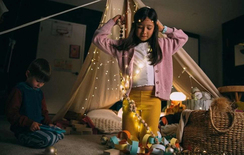 A young girl decorates a tent with string lights and wooden beads, while a young boy plays with blocks nearby indoors.