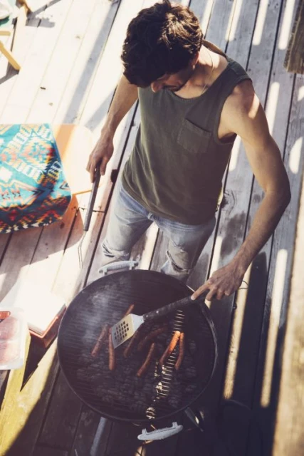 Overhead view of a man grilling hotdogs on a charcoal grill outdoors on a wooden deck, wearing a tank top and jeans.