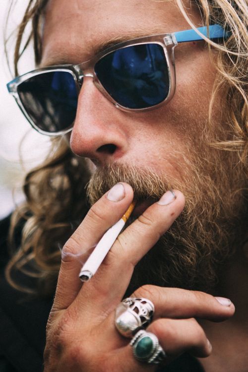 Close-up portrait of a man with long blond hair and beard wearing blue-tinted sunglasses, smoking a cigarette, with rings on his fingers.