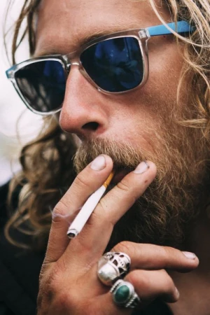 Close-up portrait of a man with long blond hair and beard wearing blue-tinted sunglasses, smoking a cigarette, with rings on his fingers.