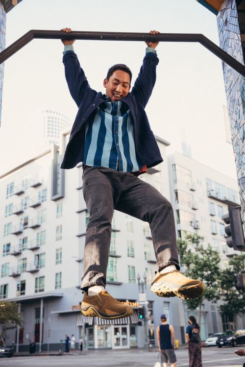 Athletic Asian man hangs from outdoor pull-up bar in an urban setting with apartment buildings, wearing dark pants and brown slip-on shoes.