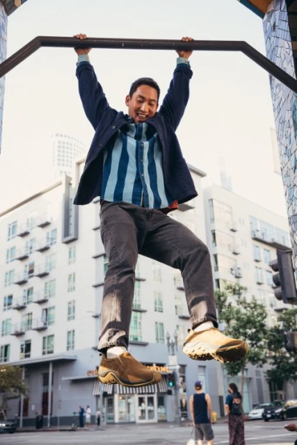 Athletic Asian man hangs from outdoor pull-up bar in an urban setting with apartment buildings, wearing dark pants and brown slip-on shoes.