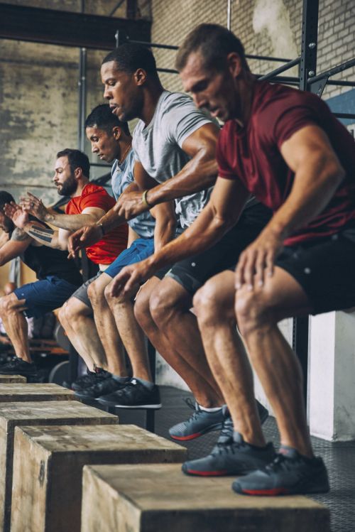 A row of adult males participate in a fitness class at an indoor gym, performing box jumps for a cross-training workout.