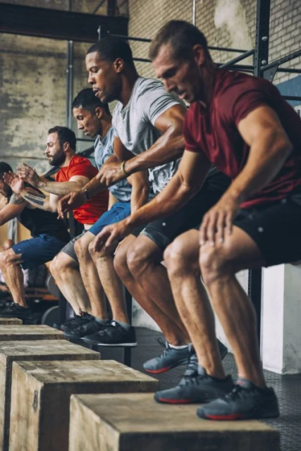 A row of adult males participate in a fitness class at an indoor gym, performing box jumps for a cross-training workout.