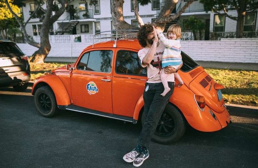 Man and toddler high-fiving next to an orange Volkswagen Beetle car on a tree-lined street with a house in the background.