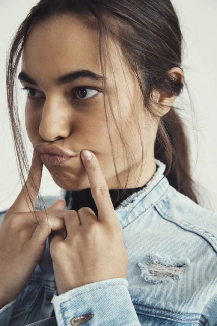 Close-up portrait of a young woman with fingers on her cheeks, wearing a denim jacket against a white background, with a playful facial expression.