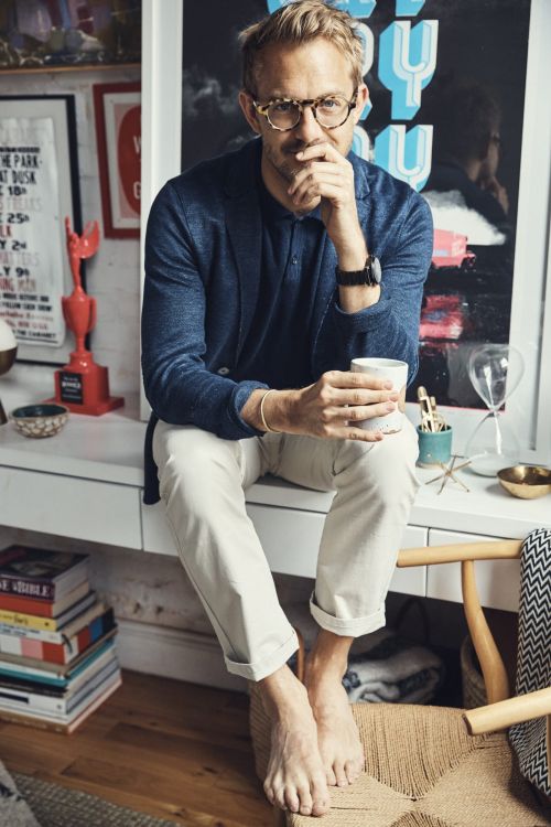 Adult male with glasses sits on a white vanity, holding a cup, with books and wall art in the background in a relaxed indoor setting.