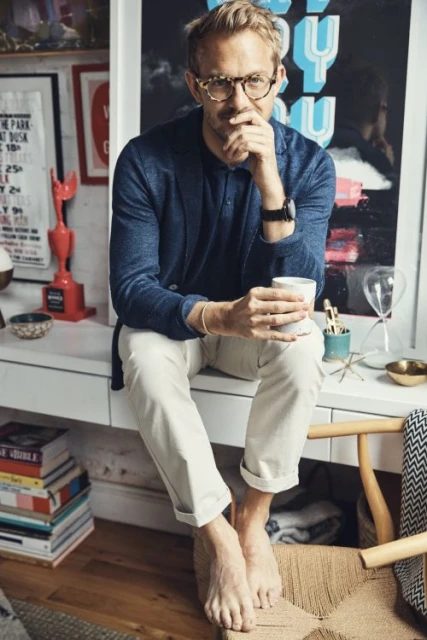 Adult male with glasses sits on a white vanity, holding a cup, with books and wall art in the background in a relaxed indoor setting.