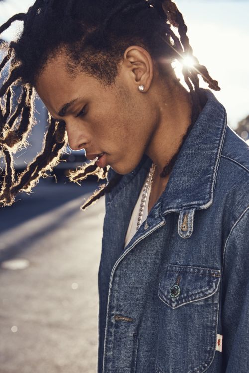 Portrait of a young adult with dreadlocks wearing a denim jacket outdoors, captured in diffused sunlight with a shallow depth of field.