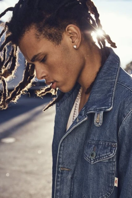 Portrait of a young adult with dreadlocks wearing a denim jacket outdoors, captured in diffused sunlight with a shallow depth of field.