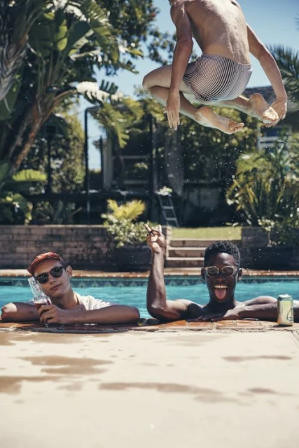 Three young men in and around an outdoor swimming pool on a sunny day. One is jumping above the water, two are in the pool, palms in background.