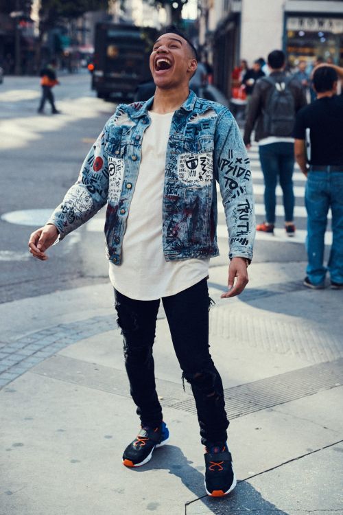 An adult male stands in an urban environment, wearing a painted denim jacket, black jeans, and tennis shoes, exuding a lively and joyful mood.