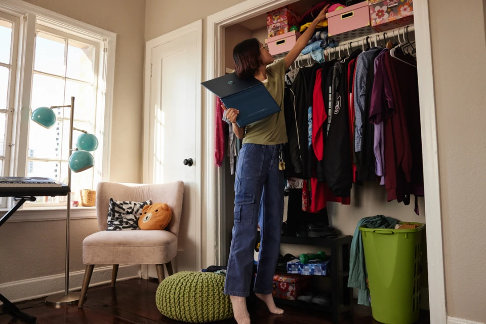 Woman standing in a bedroom organizing a closet with hanging clothes and storage boxes, laptop in hand, interior setting with lamp and chair.