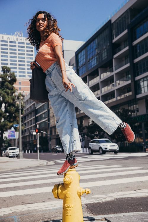 A woman balances on a yellow fire hydrant on a city street in the daytime, wearing jeans, an orange shirt, and carrying a gray tote bag.