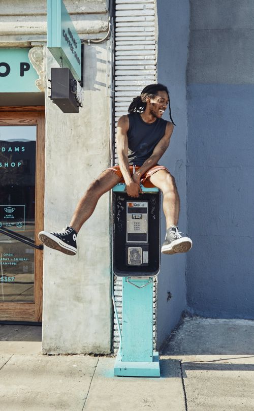 Man sits atop a vintage telephone booth in city setting, smiling with tank top, shorts, and sneakers. Outdoor shot with urban background.