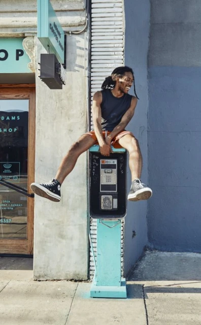 Man sits atop a vintage telephone booth in city setting, smiling with tank top, shorts, and sneakers. Outdoor shot with urban background.