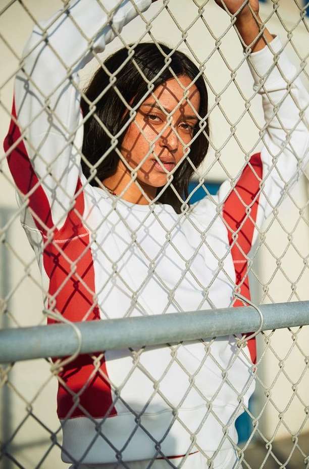 Portrait of a woman with dark hair looking through a chain-link fence, wearing a red and white sweatshirt on a sunny day.
