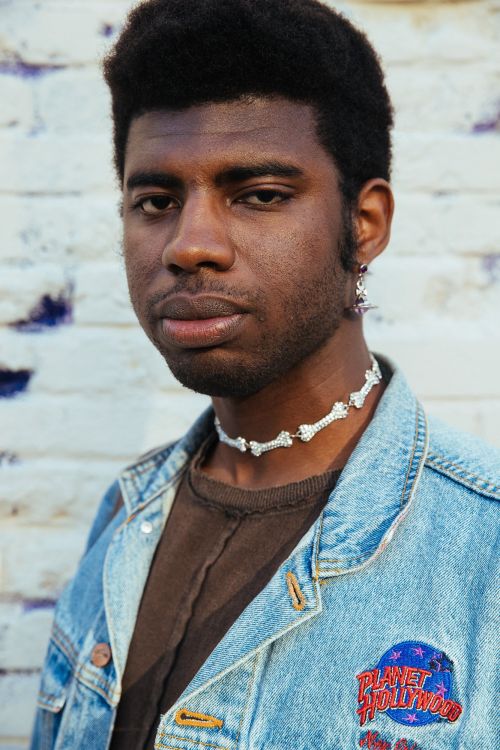 Portrait of a young Black man wearing a bone necklace and a light blue denim jacket with a Planet Hollywood patch against a brick wall.