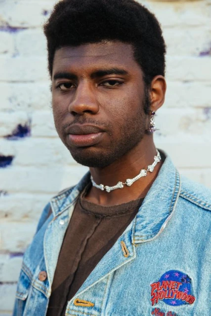Portrait of a young Black man wearing a bone necklace and a light blue denim jacket with a Planet Hollywood patch against a brick wall.