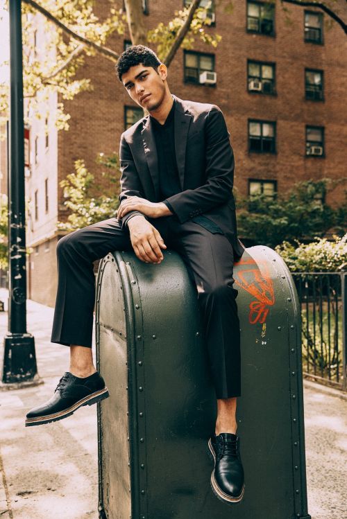 Young man in a black suit sitting on a large green post box on a New York street, wearing black shoes.