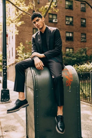 Young man in a black suit sitting on a large green post box on a New York street, wearing black shoes.