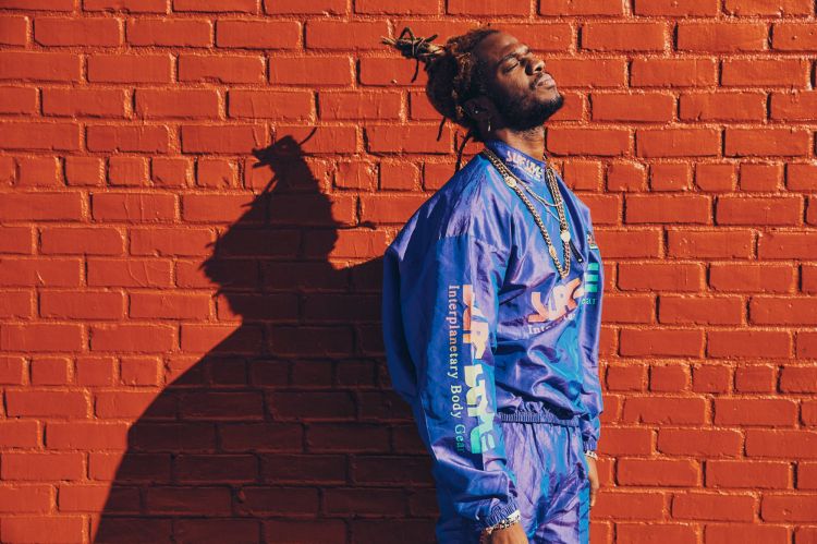 Man with dreadlocks in blue tracksuit poses against a red brick wall in sunlight, showcasing streetwear style and urban fashion.