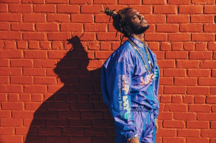 Man with dreadlocks in blue tracksuit poses against a red brick wall in sunlight, showcasing streetwear style and urban fashion.
