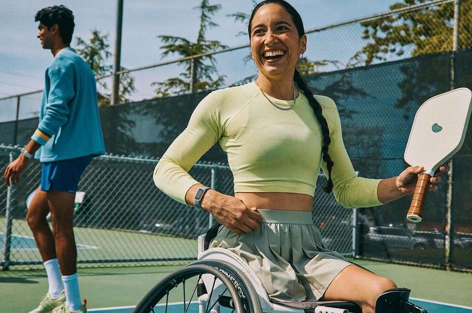 Female athlete in wheelchair smiles and holds a pickleball paddle on a tennis court with a male athlete walking in the background.