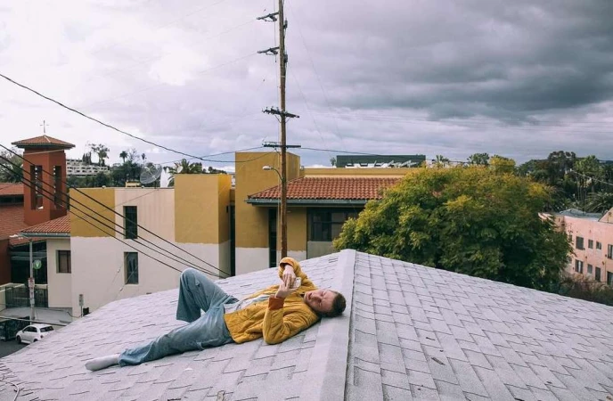 Man in yellow jacket and blue jeans lying on a shingled rooftop looking at his phone under an overcast sky.
