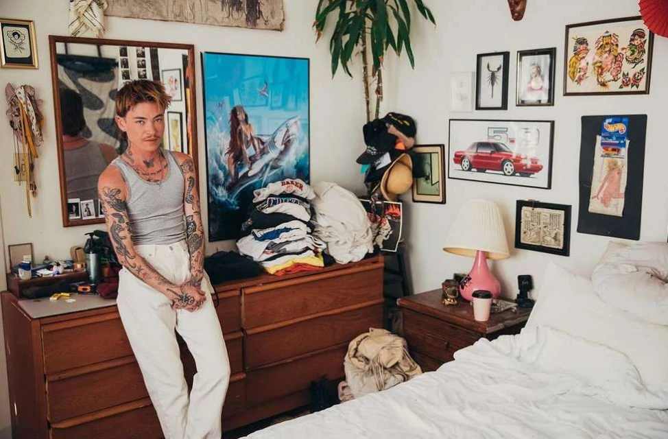 Adult male with tattoos poses in a bedroom with a dresser, bed, and framed pictures on the wall. Indoor shot, casual fashion.
