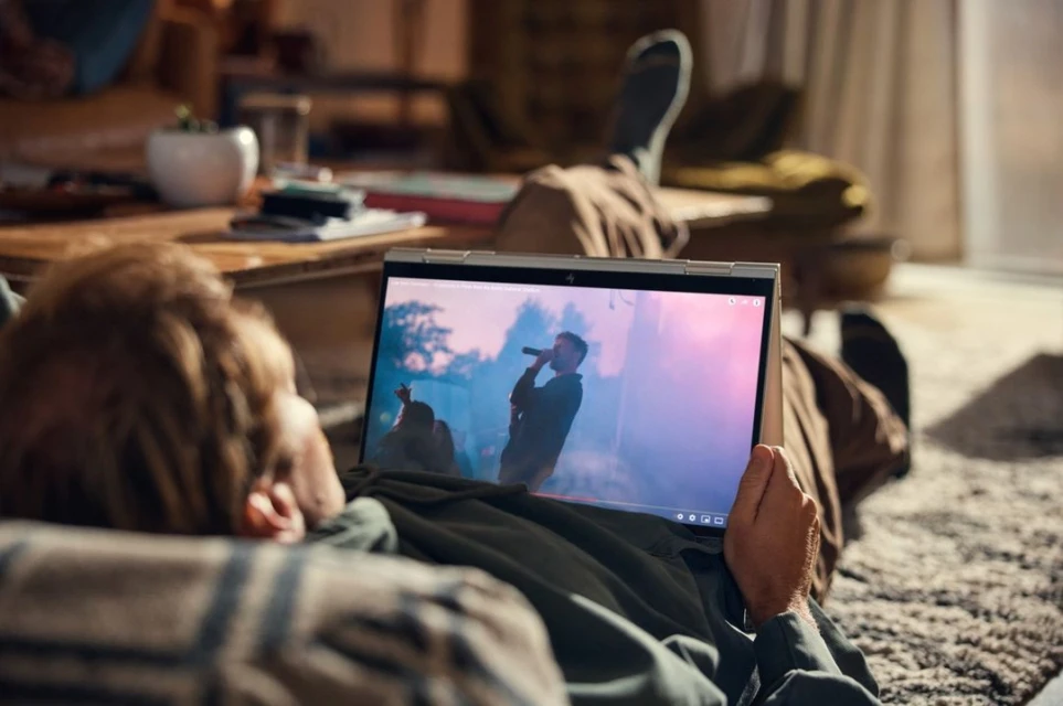 Man relaxes on a couch, watching a laptop displaying a music concert, creating a cozy, indoor scene with soft lighting.